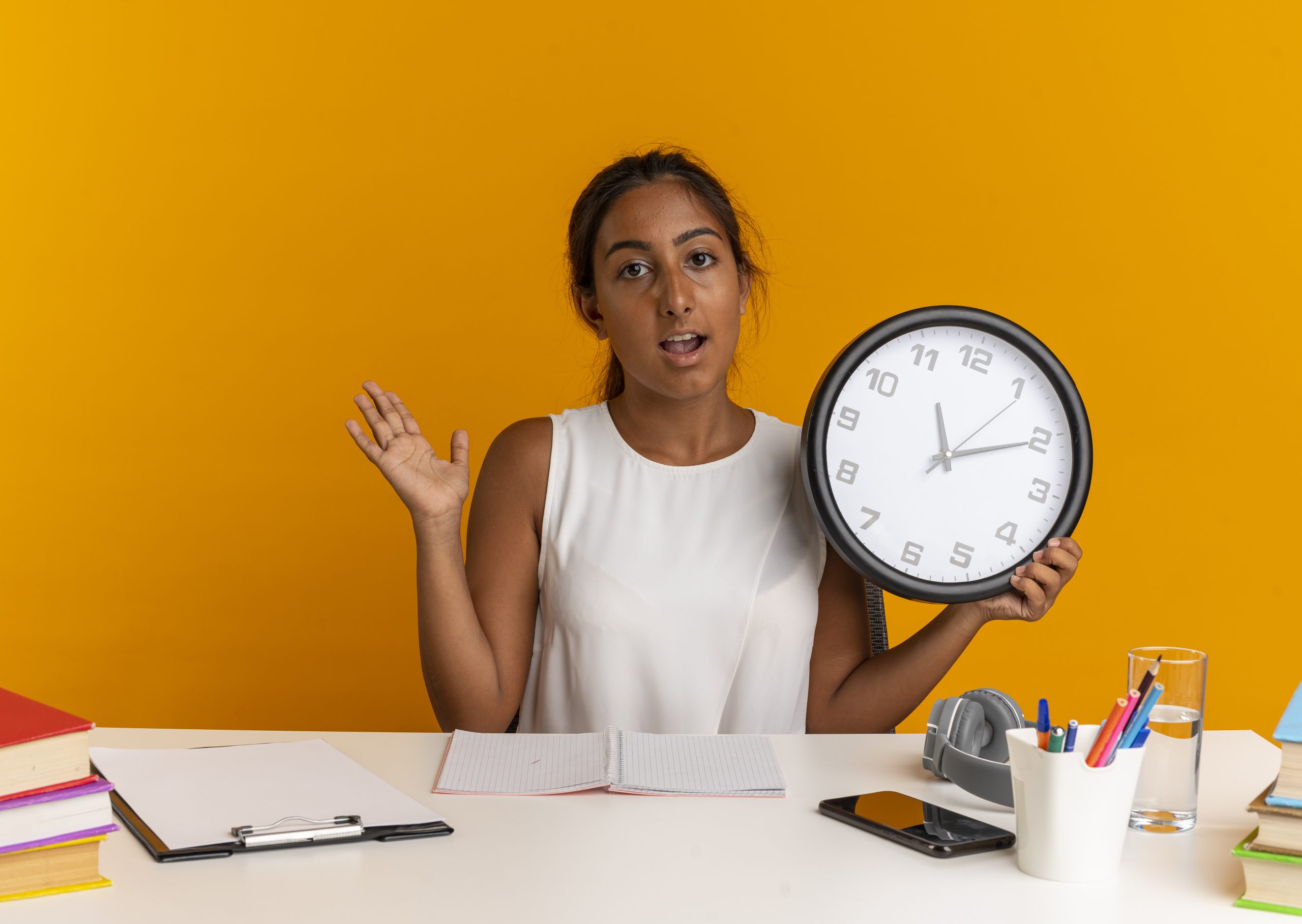pleased-young-schoolgirl-sitting-desk-with-school-tools-holding-wall-clock-spread-hand-orange-scaled.jpg